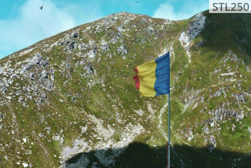 Romanian flag waves atop a scenic mountain landscape under a clear blue sky.
